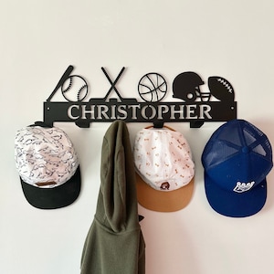 May include: Black metal sports themed coat rack with the name "CHRISTOPHER" in silver lettering. The rack has hooks for hanging hats and coats. Three hats are hanging on the hooks, one with a white and black dinosaur pattern, one with a white and brown dog bone pattern, and one blue and black mesh hat with a white logo.