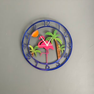 May include: A blue metal wall clock with a pink flamingo and palm trees. The clock has a white background and black numbers.