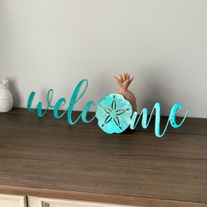 Może przedstawiać: Metalowy napis powitalny w kolorze turkusowym z piaskową monetą w środku. Napis znajduje się na drewnianej powierzchni.