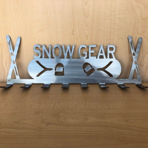 Könnte beinhalten: Metallischer Schneeausrüstungsständer mit Haken zum Aufhängen von Skiausrüstung. Der Ständer trägt die Aufschrift "SNOW GEAR" und ein stilisiertes Bild von Skiern und einem Helm.