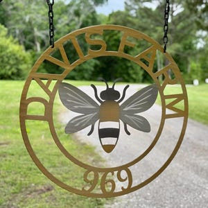 May include: A round, gold-colored metal sign with the words "DAVIS FARM" cut out around the edge. A stylized bee with gray and yellow wings and body is in the center. The number "969" is at the bottom. The sign hangs from a black chain.
