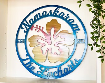 May include: A blue metal circle with the text "Namaskaram The Lochharts EST 2000" around a pink and gold hibiscus flower.