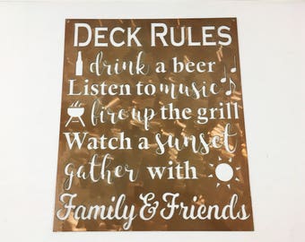 May include: A copper-colored metal sign with white text that reads "DECK RULES" followed by a list of rules: "drink a beer", "Listen to music", "fire up the grill", "Watch a sunset", "gather with", "Family & Friends".