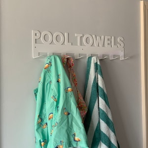 Pode incluir: Toalheiro de parede de metal branco com as palavras "POOL TOWELS" em letras grandes. O toalheiro tem 8 ganchos e est&aacute; segurando uma toalha verde com estampa de flamingos, uma toalha estampada em rosa e branco e uma toalha listrada verde e branca.