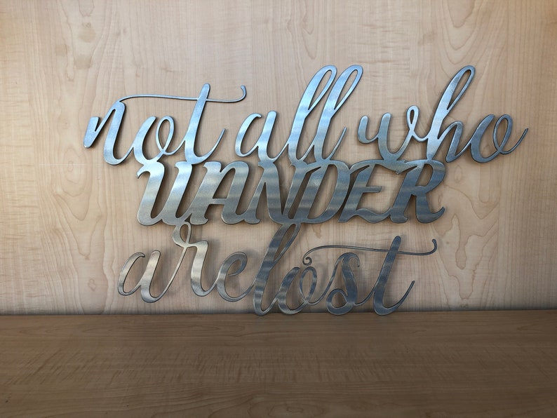 May include: Metal wall art with the quote "Not all who wander are lost". The quote is written in a cursive font and is made of silver metal.