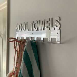 Pode incluir: Um suporte de toalha de metal branco com as palavras "POOL TOWELS" recortadas. O suporte tem seis ganchos e uma toalha e um mai&ocirc; est&atilde;o pendurados nos ganchos.