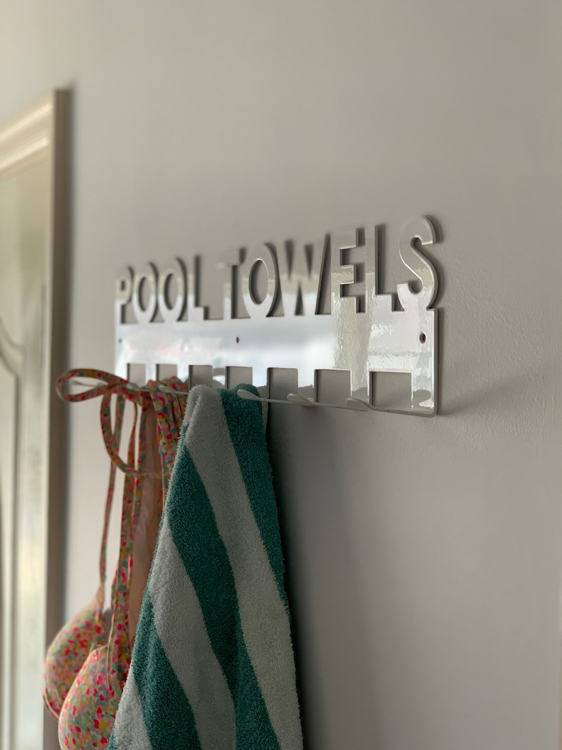 Pode incluir: Um suporte de toalha de metal branco com as palavras "POOL TOWELS" recortadas. O suporte tem seis ganchos e uma toalha e um mai&ocirc; est&atilde;o pendurados nos ganchos.