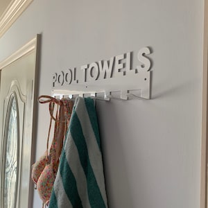 Pode incluir: Um suporte de toalha de parede de metal branco com as palavras "POOL TOWELS" em letras brancas. Uma toalha listrada verde e branca e um biqu&iacute;ni com estampa floral est&atilde;o pendurados no suporte.