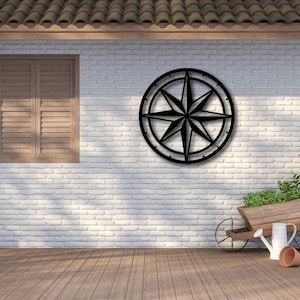May include: A black metal compass rose wall art is mounted on a white brick wall. A wooden shuttered window is to the left. A wooden wheelbarrow with plants, gardening tools, and boots are in front.