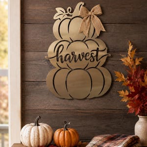 May include: A gold-toned metal wall decoration shaped like a stack of three pumpkins with the word harvest cut out in cursive script. The top pumpkin features a decorative burlap bow attached to the side, and the entire piece has a smooth finish.