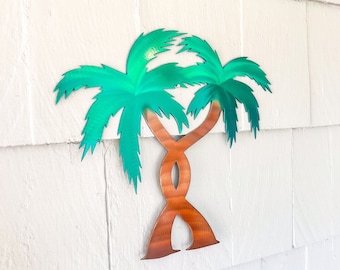 May include: A metal wall decor featuring two intertwined palm trees with green leaves and a brown trunk. The palm trees are against a white background.