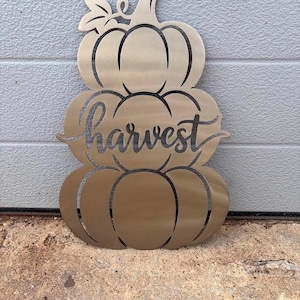 May include: A metallic gold-toned wall decoration shaped like a stack of three pumpkins. The center pumpkin features the word harvest in a cursive script font. The design includes a stem at the top and a single leaf detail on the left side.