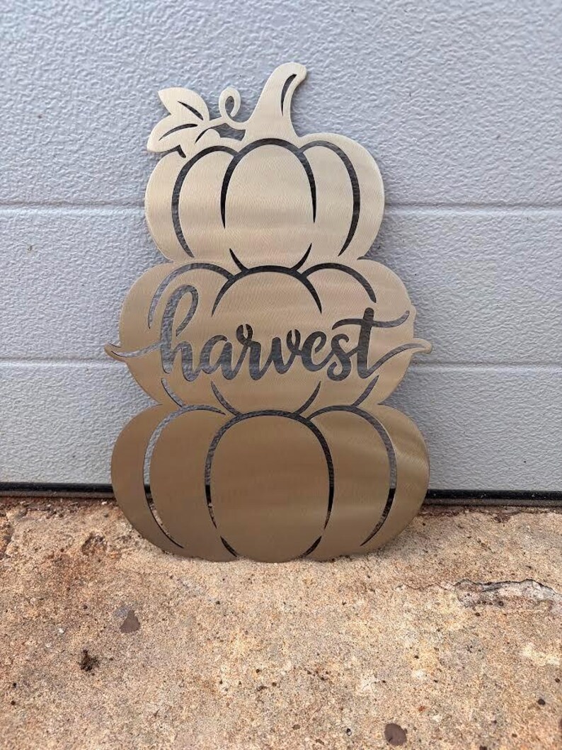 May include: A metallic gold-toned wall decoration shaped like a stack of three pumpkins. The center pumpkin features the word harvest in a cursive script font. The design includes a stem at the top and a single leaf detail on the left side.