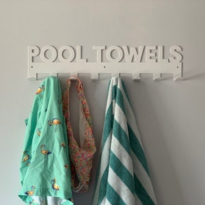 Pode incluir: Toalheiro de parede de metal branco com as palavras "POOL TOWELS" em letras grandes. O toalheiro tem sete ganchos e est&aacute; segurando uma toalha verde com estampa de flamingos, uma toalha estampada em rosa e branco e uma toalha listrada azul-esverdeada e branca.