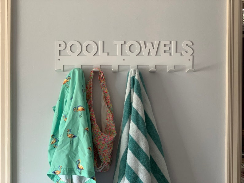 Pode incluir: Toalheiro de parede de metal branco com as palavras "POOL TOWELS" em letras grandes. O toalheiro tem sete ganchos e est&aacute; segurando uma toalha verde com estampa de flamingos, uma toalha estampada em rosa e branco e uma toalha listrada azul-esverdeada e branca.