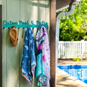 May include: A teal wooden sign with the text "Custom Pool & Patio" hangs on a light green wooden wall.  A straw hat, a blue towel with a shark pattern, and a colorful patterned towel hang on hooks attached to the sign.