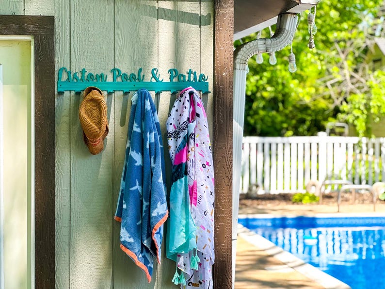 May include: A teal wooden sign with the text "Custom Pool & Patio" hangs on a light green wooden wall.  A straw hat, a blue towel with a shark pattern, and a colorful patterned towel hang on hooks attached to the sign.