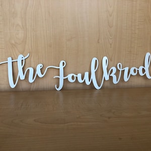 Könnte beinhalten: Ein weißes Holzschild mit dem Text "The Foulkroods" in einer kursivschrift.