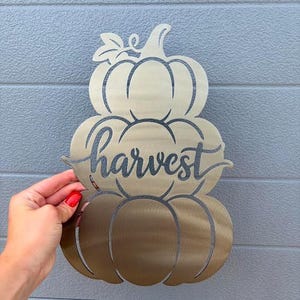 May include: A metallic gold-toned wall decoration shaped like a stack of three pumpkins. The word harvest is written in a cursive script across the middle pumpkin. The piece is constructed from a flat sheet of metal with cut-out details.