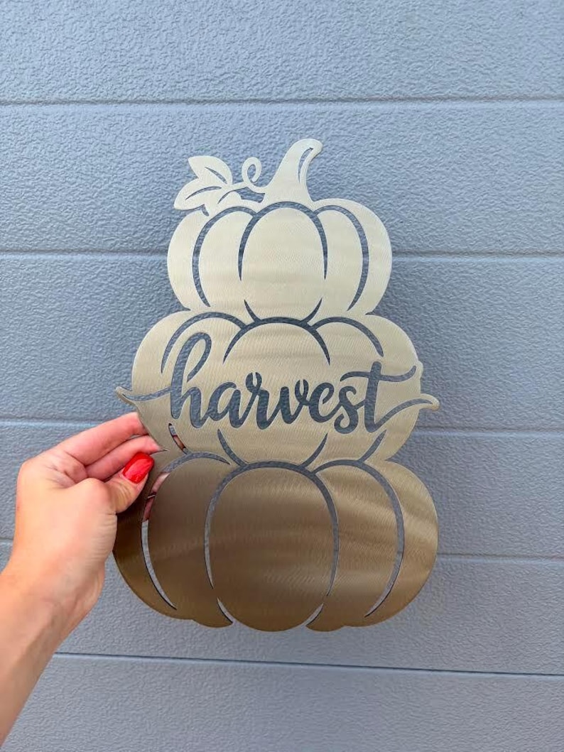 May include: A metallic gold-toned wall decoration shaped like a stack of three pumpkins. The word harvest is written in a cursive script across the middle pumpkin. The piece is constructed from a flat sheet of metal with cut-out details.