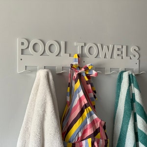 Pode incluir: Um suporte de toalha de parede de metal branco com as palavras "POOL TOWELS" impressas nele. Tr&ecirc;s toalhas est&atilde;o penduradas no suporte, uma branca, uma listrada verde e branca e uma listrada rosa, amarela e preta.