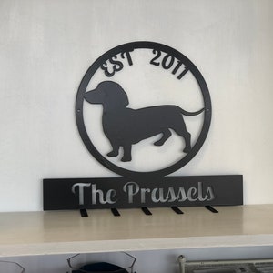 May include: Black metal wall decor featuring a dachshund silhouette inside a circle with the text "EST 2011". Below the circle is a black metal sign with the text "The Prassels" and five hooks.