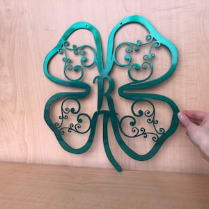 May include: A green metal four-leaf clover with a decorative swirl design and the letter "R" in the center.