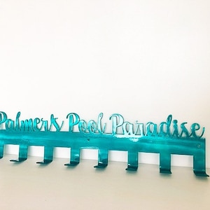 May include: Teal metal wall hook with the text "Palmers Pool Paradise" and eight hooks.