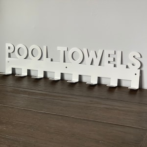 Pode incluir: Toalheiro de metal branco com as palavras "POOL TOWELS" em letras grandes. O toalheiro tem 10 ganchos para pendurar toalhas.