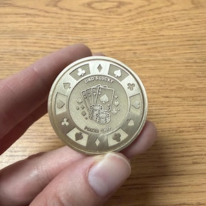 May include: A gold-coloured poker chip with the text "Dad's Lucky" and "Poker Chip" engraved on it. The chip features card suit symbols and a poker hand design. The chip is round and has a detailed, textured surface.