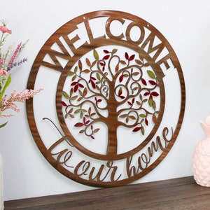Puede incluir: Un letrero redondo de madera con un diseño de árbol y las palabras "Bienvenido a nuestro hogar" en una fuente decorativa. El letrero está colgado en una pared blanca y hay flores en un jarrón y dos recipientes con forma de piña sobre una superficie de madera debajo.
