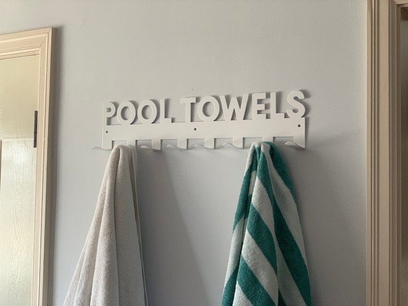 Pode incluir: Um suporte de toalha de metal branco com as palavras "POOL TOWELS" nele. H&aacute; duas toalhas penduradas no suporte, uma branca e outra verde e branca listrada.