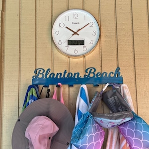 May include: A blue metal sign with the words "Blanton Beach" on it. The sign has hooks for hanging towels and other items. There are towels, a hat, and a life vest hanging on the hooks. A clock with a white face and gold trim is mounted on the wall behind the sign.