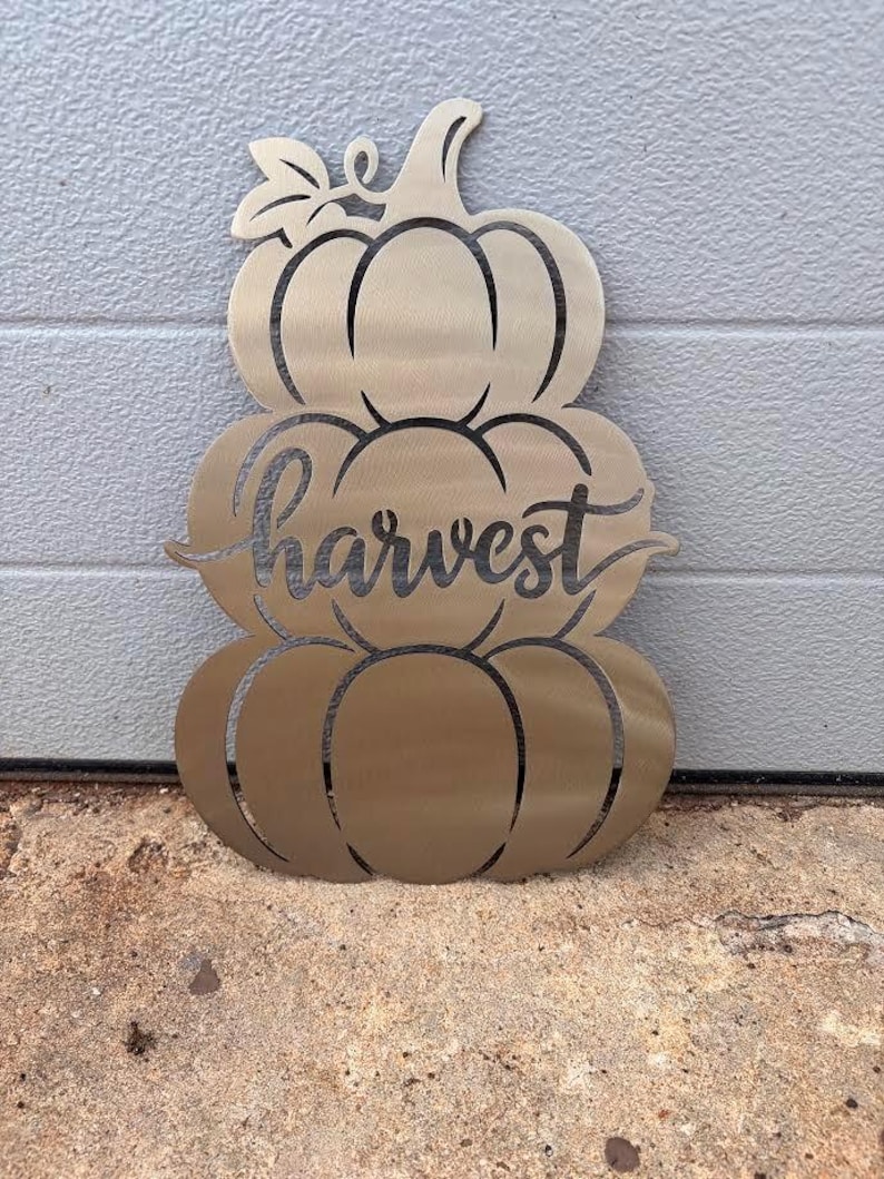 May include: A metallic gold-toned wall decoration shaped like a stack of three pumpkins. The center pumpkin features the word harvest in a cursive script font. The design includes a stem at the top and a single leaf detail on the left side.