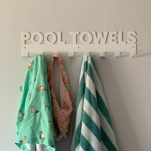 Pode incluir: Um suporte de parede de metal branco com as palavras "POOL TOWELS" em letras recortadas. O suporte tem oito ganchos e est&aacute; segurando uma toalha verde com estampa de flamingo, uma toalha estampada multicolorida e uma toalha listrada verde e branca.