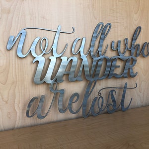 May include: Metal wall art with the quote "Not all who wander are lost". The quote is written in a cursive font and is made of brushed metal.