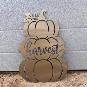 May include: A gold-toned metal wall decoration shaped like a stack of three pumpkins. The design features cut-out lines defining the pumpkin segments and a leaf at the top. The word harvest is written in a cursive script across the middle pumpkin.