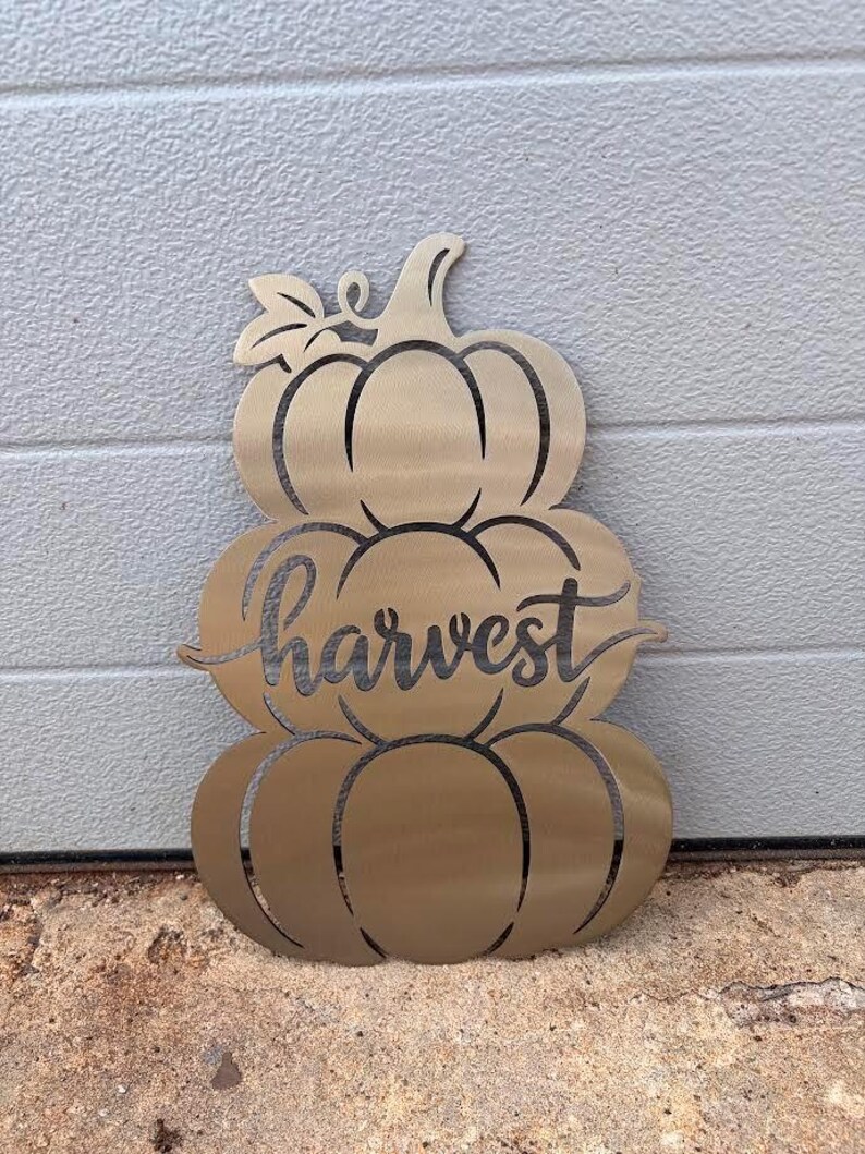 May include: A gold-toned metal wall decoration shaped like a stack of three pumpkins. The design features cut-out lines defining the pumpkin segments and a leaf at the top. The word harvest is written in a cursive script across the middle pumpkin.