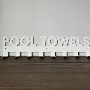 Pode incluir: Toalheiro de parede de metal branco com as palavras "POOL TOWELS" recortadas. O toalheiro tem oito ganchos.