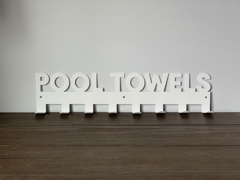 Pode incluir: Toalheiro de parede de metal branco com as palavras "POOL TOWELS" recortadas. O toalheiro tem oito ganchos.