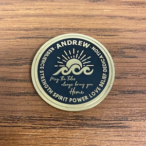 May include: A gold-colored round coin with a black center. The coin has the words "ANDREW" and "RESILIENCE STRENGTH SPIRIT POWER LOVE BELIEF DEDICATION" around the edge. The center of the coin has a sun with rays and waves. The words "May the tides always bring you Home" are written below the sun and waves.
