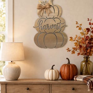May include: A gold-toned metal wall hanging shaped like a stack of three pumpkins with the word harvest cut out in cursive script across the center. A tan burlap bow is attached to the top pumpkin stem, adding a textured accent to the piece.