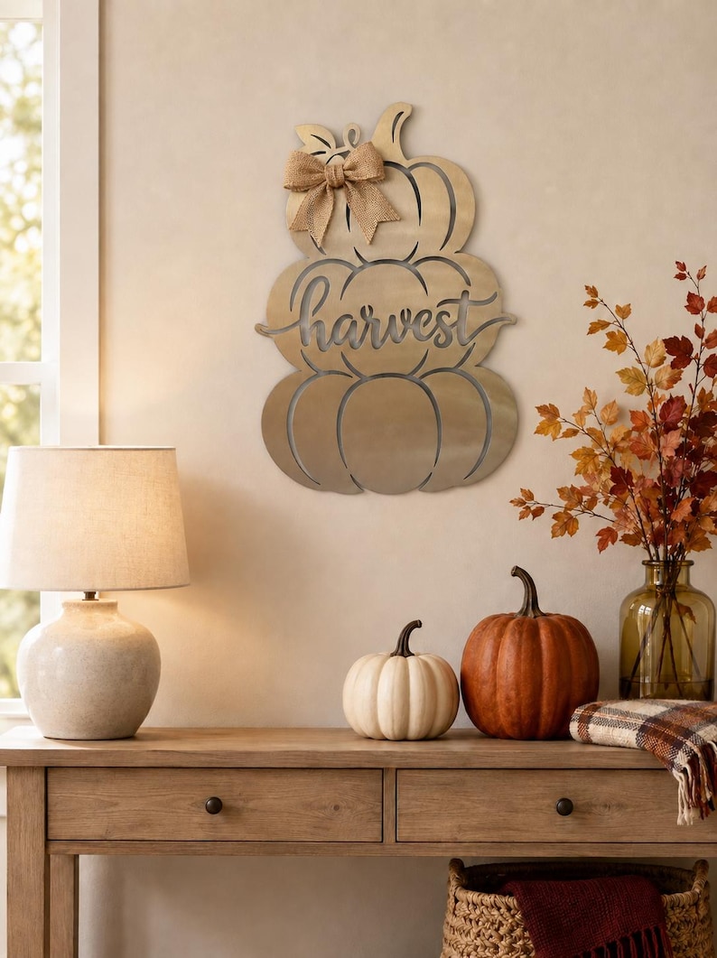 May include: A gold-toned metal wall hanging shaped like a stack of three pumpkins with the word harvest cut out in cursive script across the center. A tan burlap bow is attached to the top pumpkin stem, adding a textured accent to the piece.