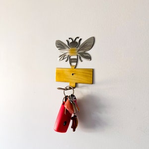 May include: A metal bee-shaped key holder with a yellow rectangular base. A set of keys is hanging from the hook.