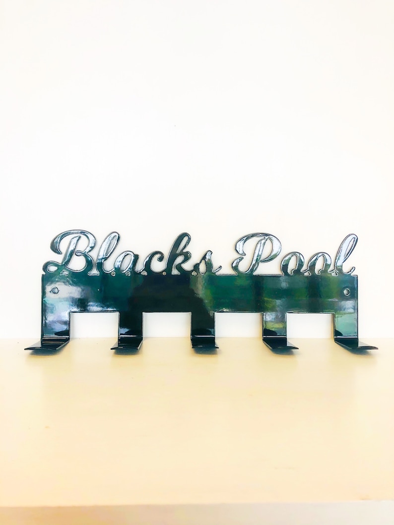 May include: Black metal wall hook with the words "Blacks Pool" in a decorative font. The hook has five hooks for hanging items.