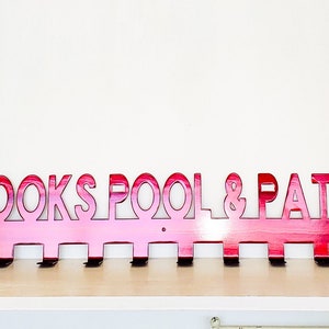 May include: A pink metal coat rack with the words "COOKS POOL & PATIO" cut out. The rack has eight hooks.