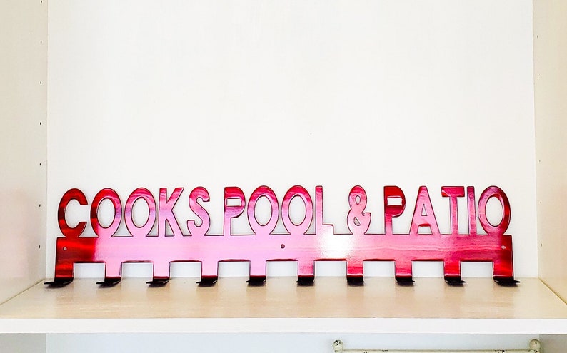 May include: A pink metal coat rack with the words "COOKS POOL & PATIO" cut out. The rack has eight hooks.