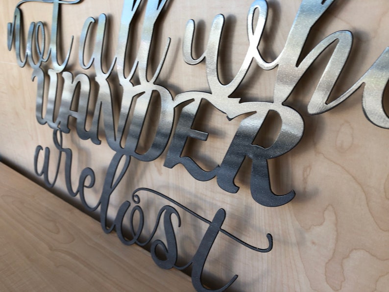 May include: Metal wall art with the quote "Not all who wander are lost". The art is made of brushed metal and has a modern, minimalist style.