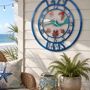 May include: A blue metal wall clock with a mermaid design. The clock features a circular frame with Roman numerals and the words "LAZY DAYS". The mermaid has a white body and a teal tail, with coral accents.
