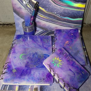 May include: Four purple and white galaxy themed notebooks with silver glitter and holographic accents. The notebooks have different designs including a crescent moon, a sun, a pentagram, and a hand with a planet. The notebooks are bound with silver rings.